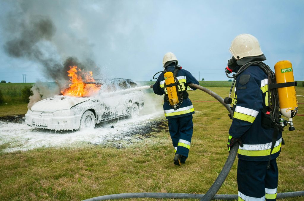 Firefighters tackle a blazing car using foam to extinguish the flames in an outdoor setting.