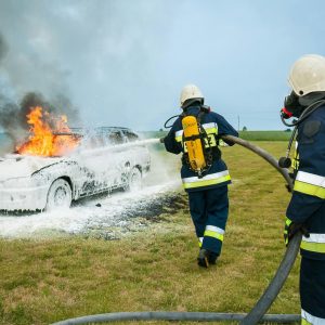 Firefighters tackle a blazing car using foam to extinguish the flames in an outdoor setting.